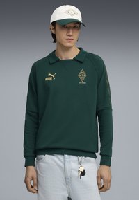 Tânăr îmbrăcat într-un hanorac verde închis Puma cu logo-urile "KING" și Borussia Mönchengladbach 125 Jahre, șapcă albă și blugi deschiși, având un breloc.
