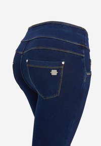 Tummansiniset korkeavyötäröiset farkut, joissa on takataskussa metallisia neppareita ja brändätty neliön muotoinen metallinen tunniste yksivärisellä taustalla.