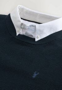 Maglione a coste navy indossato sopra una camicia bianca con colletto a pulsante, dotata di un piccolo logo ricamato di un cervo sul petto.