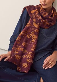 Écharpe à motifs bordeaux et or avec une texture côtelée, portée lâchement autour du cou, associée à une tenue en denim.