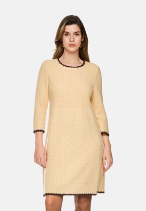 Woman wearing a beige textured long-sleeve dress with dark brown trim on the neckline, cuffs, and hem, standing against a white background.