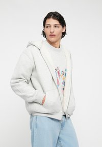 Un sweat à capuche zippé gris clair avec une capuche en sherpa et des poches frontales, superposé à un sweatshirt à motif graphique et associé à un jean bleu clair.