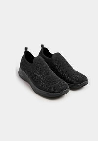 Chaussures noires à enfiler en matériau tricoté, présentant une surface texturée avec des accents scintillants et une semelle noire rembourrée.
