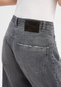 Jean en denim gris avec une coupe décontractée, des détails usés et une patch en cuir à la taille arrière. Comprend des poches arrière avec des coutures.