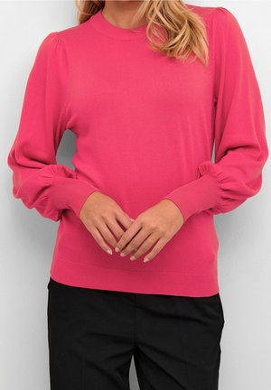 Femme portant un pull rose vif à manches longues avec des manches bouffantes et un pantalon noir, debout les mains légèrement croisées.