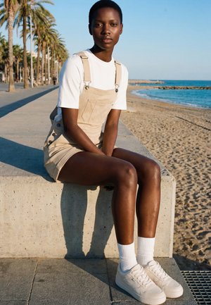 Lühikeste juustega noor inimene, kandes beeže kombinesooni ja valgeid tossusid, istumas betoonseinal ranna lähedal, kus on palmipuud ja ookean.