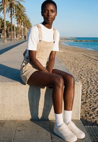 Persona joven con pelo corto que lleva un mono beige y zapatillas blancas, sentada en un muro de hormigón junto a la playa con palmeras y el océano cerca.