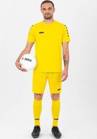 Gelbes Sportoutfit von Jako, mit kurzen Ärmeln, schwarzen Akzenten sowie passenden Shorts und Socken. Spieler hält einen weißen Jako Fußball.