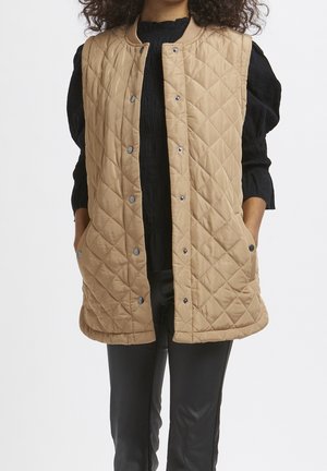 Femme portant une veste matelassée beige sans manches avec des boutons-pression sur un haut noir à manches longues et un pantalon noir, les mains dans les poches de la veste.