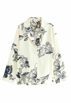 Person siddende på en træstol iført sorte bukser med vide ben og en cremefarvet blazer med store blomsterprint, kombineret med hvide sandaler med stropper.