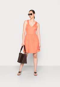 Vestido cor-de-rosa sem mangas com decote em V e cintura franzida. A modelo segura uma bolsa texturizada castanho-escura, usa óculos de sol oversized e sandálias planas.