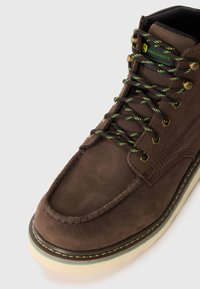 Bottines de travail en cuir marron avec des œillets de lacets verts et jaunes, estampillées du logo "John Deere", et une semelle couleur crème avec une bande verte.