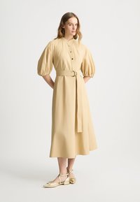 Beiges Hemdblusenkleid mit geschoppten Ärmeln, tailliertem Bund und fließendem Rock. Kombiniert mit goldenen Ballerinas mit Knöchelriemen.