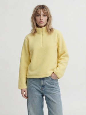 Frau steht mit der Hand in der Tasche, trägt einen gelben Fleecepullover mit halbem Reißverschluss und hellblaue Jeans vor einem schlichten Hintergrund.