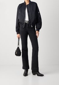 Černá bomber bunda s žebrovanými manžetami, bílý top, černé džíny s vysokým pasem a černé kotníkové boty na podpatku, držící malou černou kabelku.