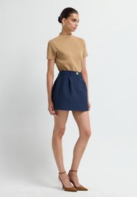 Top beige a maniche corte abbinato a una gonna plissettata in navy con un bottone dorato. Modella con tacchi alla caviglia marroni.