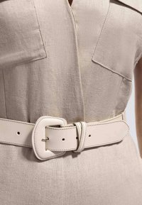 Bež lanena haljina sa širokim prednjim džepovima i odgovarajućim širokim bež remenom sa zaobljenom pravokutnom kopčom oko struka.