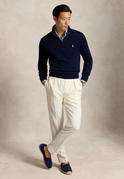 Homme portant un pull marine à fermeture éclair quart de zip, chemise rayée, pantalon plissé crème, espadrilles marine et montre-bracelet, debout les mains dans les poches.