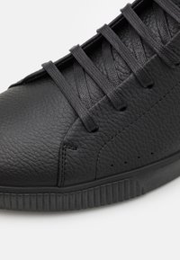 Černý kožený teniska s texturovaným povrchem, plochými tkaničkami a gumovou podrážkou s rýhovanými detaily a malými průduchy.