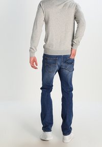 Grauer Pullover mit gerippten Bündchen, kombiniert mit blauen Jeans mit hinteren Taschen. Das Modell trägt weiße Sneakers und steht mit dem Rücken zur Kamera.