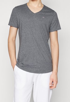 Homme portant un t-shirt gris à col en V et manches courtes et un pantalon blanc, une main dans la poche, debout devant un fond uni.