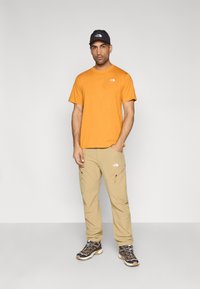 Πορτοκαλί μπλουζάκι με λογότυπο, μπεζ cargo παντελόνι με τσεπάκια με φερμουάρ, και μαύρο καπέλο. Τα παπούτσια έχουν σκούρα και ανοιχτά σχέδια με κορδόνια.