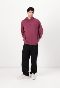 Felpa oversize color bordeaux con ricamo frontale, abbinata a pantaloni cargo neri e sneakers bianche. Tessuto liscio con polsini a coste.