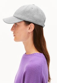 Femme aux longs cheveux bruns portant une casquette de baseball gris clair et un pull en tricot violet, montrée de profil sur fond blanc.