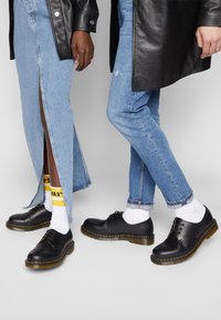 Pantofi din piele neagră cu vârful rotund, cusături galbene și talpă groasă. Purtați cu blugi din denim și șosete contrastante cu text.
