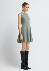 Zelené bezrukávové šaty s rozšířenou sukní, vyrobené z hladkého materiálu. V kombinaci s vysokými černými koženými botami z krokodýlí kůže a velkými náušnicemi.