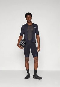 Svart cykeloutfit med en åtsittande kortärmad tröja med dragkedja och matchande shorts. En hjälm hålls i vänster hand.