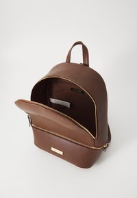 Sac à dos en cuir marron de forme arrondie, avec une fermeture éclair dorée et une poche avant. L'intérieur est doublé d'un tissu plus clair.