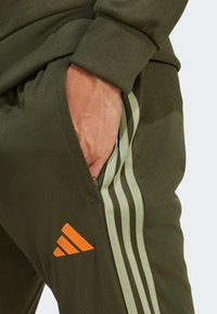 Olive green joggers with light green side stripes and an orange Adidas logo patch. Features a zippered pocket and ribbed cuffs.
