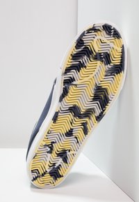Suola di una sneaker blu con un modello di battistrada zigzag multicolore in giallo, bianco e nero, poggiata su una superficie bianca.