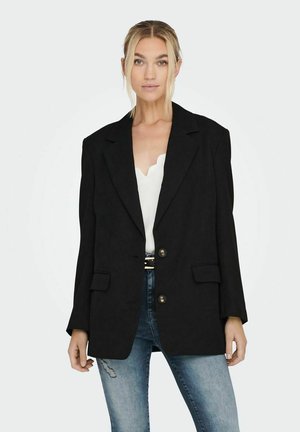 Blazer nero con un design strutturato, chiusura a un solo bottone, spalline prominenti e tasche frontali. Indossato sopra una maglietta bianca.