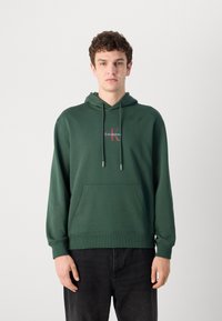 Felpa con cappuccio verde, dotata di tasca frontale, polsini e orlo a coste. Presenta un logo rosso e bianco sul petto. Il materiale sembra morbido.