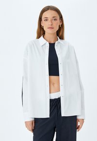 Witte lange mouwen knoopshirt met een relaxte pasvorm, subtiele glans en een klassieke kraag, gecombineerd met een zwarte crop top en donkere broek.