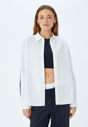 Witte lange mouwen knoopshirt met een relaxte pasvorm, subtiele glans en een klassieke kraag, gecombineerd met een zwarte crop top en donkere broek.
