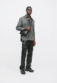 Un tânăr îmbrăcat cu o cămașă gri cu nasturi, pantaloni lungi din piele cargo negri, adidași negri, ținând o geantă neagră de umăr pe un fundal alb.