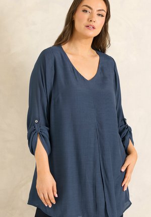 Mujer con blusa azul marino suelta de manga larga, cuello en V y lazos abotonados en las mangas, de pie contra un fondo claro y liso.