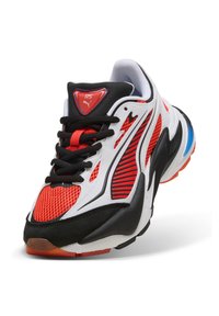 Zapatilla deportiva con malla roja, superposiciones en blanco y negro, cordones negros y un detalle azul en la suela, con el logo de Puma RS en la lengüeta.