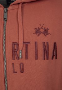 Roestkleurige zip-up hoodie van textuurstof, met geborduurde letters en een paardenafbeelding. Zilveren ritsaccent.
