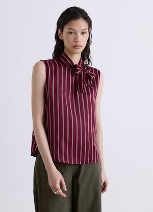 Giovane donna che indossa una camicetta senza maniche color granata con strisce bianche verticali e un fiocco al collo, abbinata a pantaloni verde oliva.