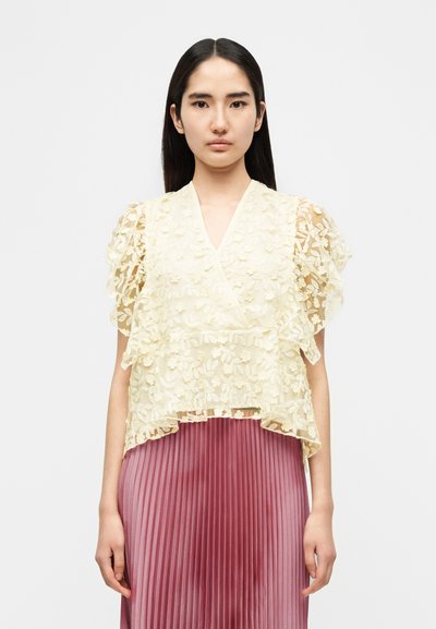 Κίτρινη μπλούζα από διαφανές δαντελωτό ύφασμα με κοντά μανίκια με βολάν και ντεκολτέ σε σχήμα V, συνδυασμένη με πτυχωτή ροζ φούστα, δημιουργώντας οπτική αντίθεση υφής.