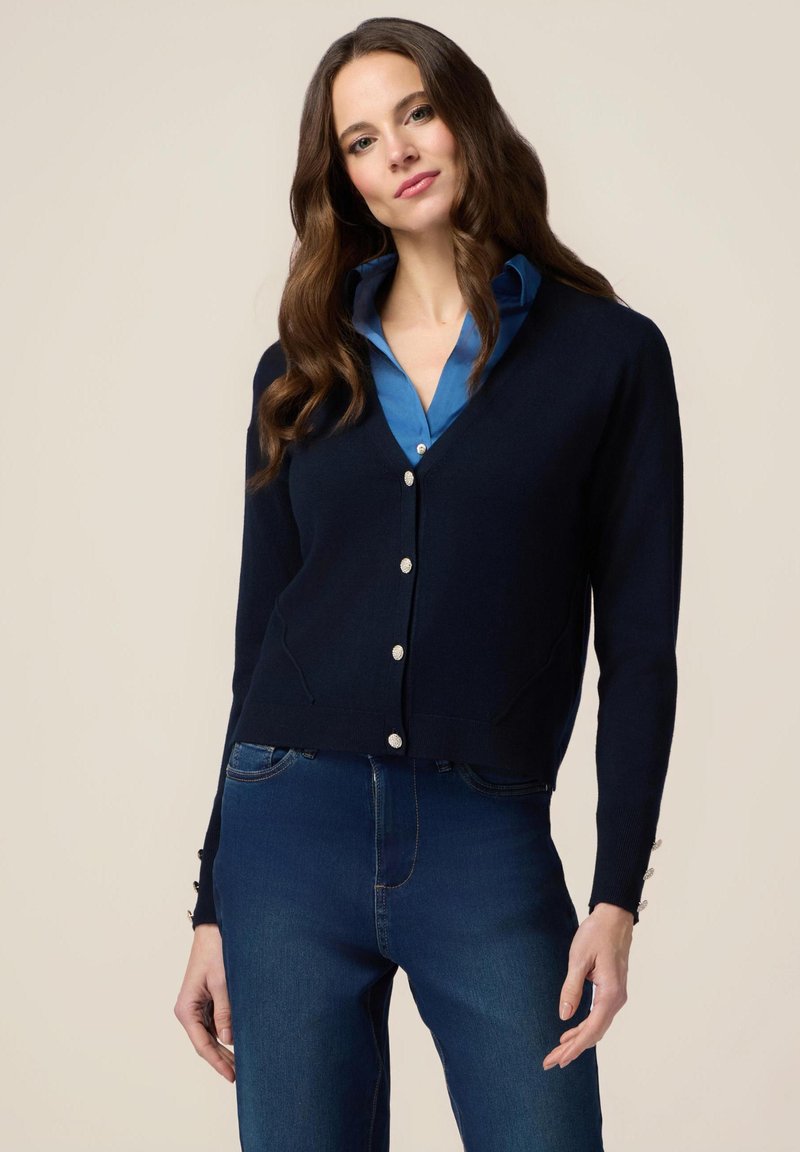 Cardigan bleumarin cu decolteu în V, nasturi argintii și buzunare laterale. Purtați peste o cămașă albastră, asortat cu blugi. Textură netedă, design ajustat.