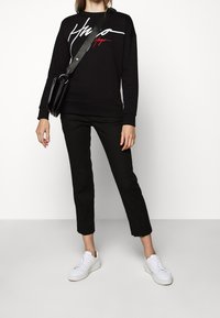 Sweat à capuche noir avec du texte blanc et rouge sur le devant. Associé à un pantalon court noir et des baskets blanches. Mannequin tenant un sac noir.