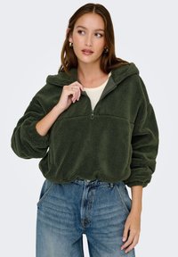 Un pulóver de forro polar verde oliva, con cremallera frontal y capucha. Presenta una textura suave, mangas largas y un corte ligeramente recortado. Combinado con jeans azules.