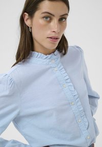 Chemise bleu clair avec col à volants, boutonnage devant et manches bouffantes. Fabriquée en tissu doux avec une texture subtile et une coupe ajustée.