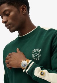 Sudadera verde con acentos crema y logo, con puños acanalados. Reloj dorado con esfera azul y correa de cuero en la muñeca.