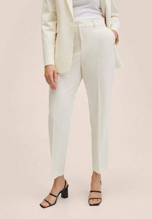 Femme portant un pantalon crème sur mesure, un blazer assorti, un haut côtelé blanc et des sandales à talons noires, debout avec une main dans la poche.